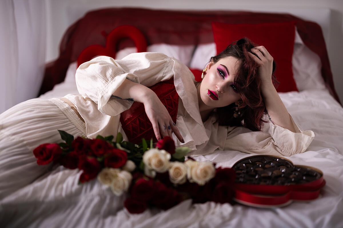 Woman wearing a red corset and white blouse reclining on a bed with red roses and a heart-shaped box of chocolates.